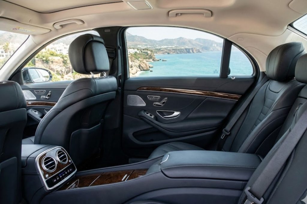 Interior de coche de lujo con asientos de cuero negro y detalles en madera, vista panorámica al mar.