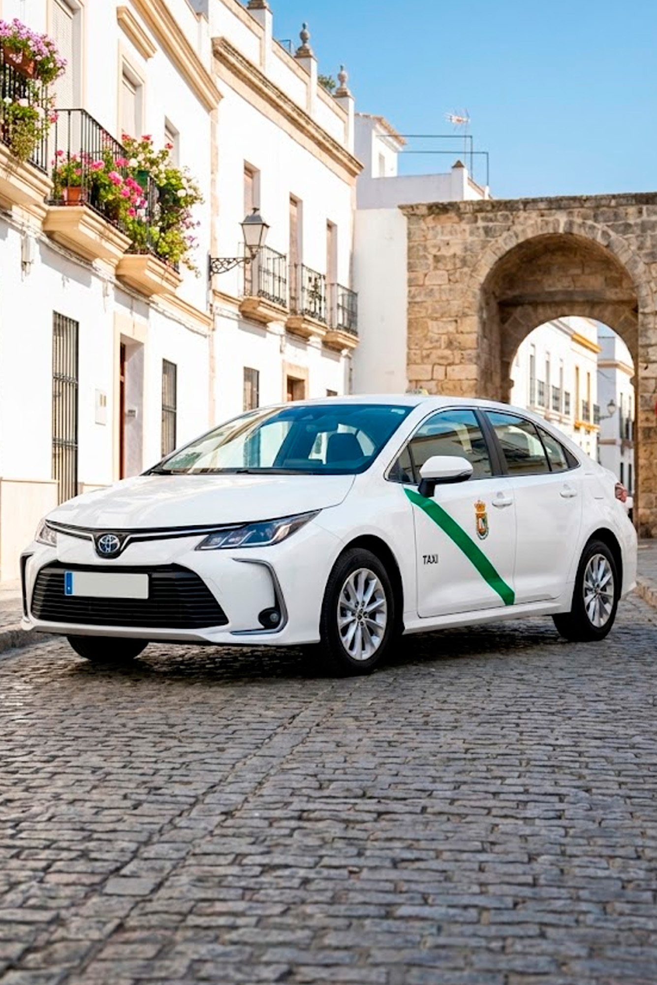 Taxi Toyota Corolla blanco estacionado en una calle empedrada con edificios históricos y arco de piedra al fondo en España.